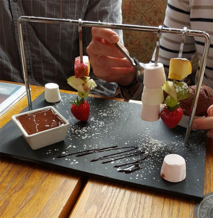 Creative restaurant food serving with skewered fruits and marshmallows on a slate, featuring a chocolate dip.
