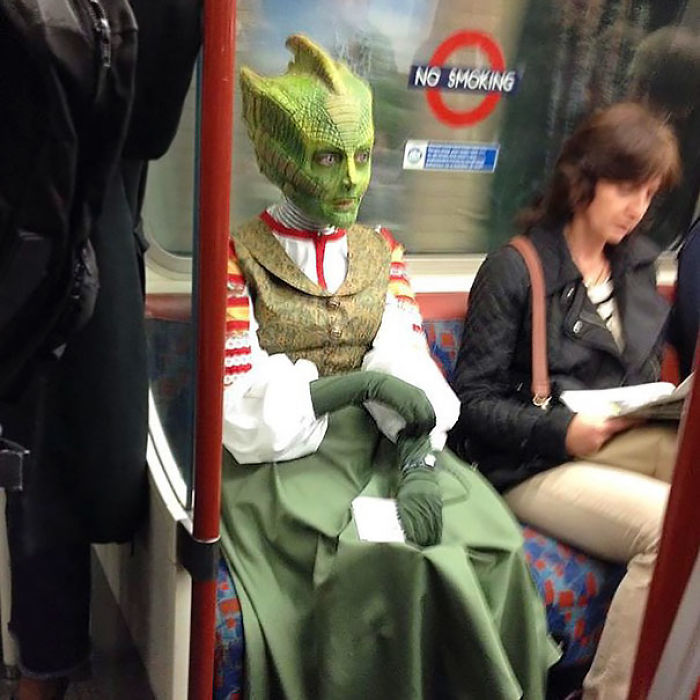 Person in a green alien costume sitting on the subway, surrounded by commuters. Weird subway sightings.