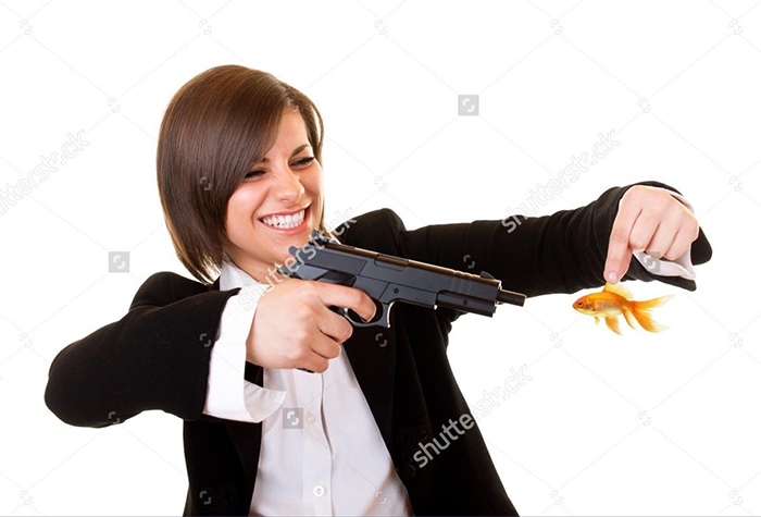 Happy woman in a classy outfit in one hand holding a gun and in other goldfish - preparing to shoot it