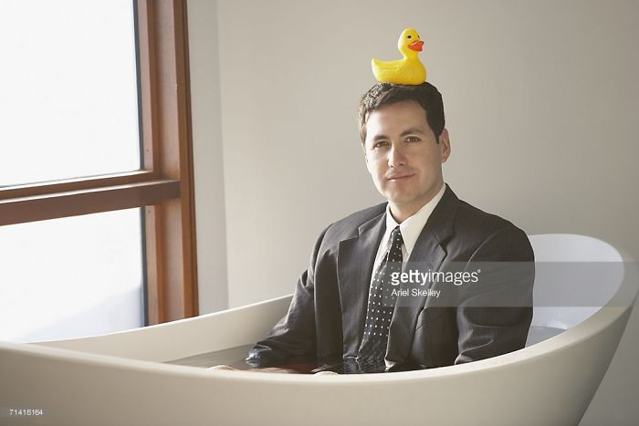 A man in a suit sitting in a tub and a yellow rubber duck is on his head