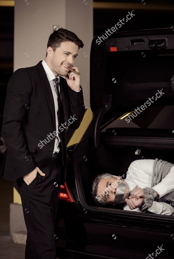 A man is casually smiling while taking a smoke break and talking on the phone, and there is a man tied up in his trunk