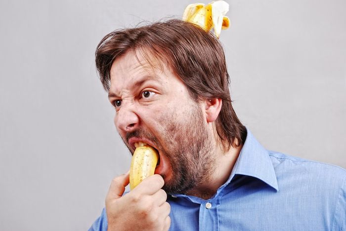 A man in a blue shirt stabs himself with a banana which goes through his mouth and upper head