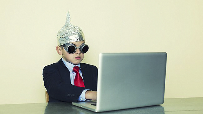 A boy in a suit, sunglasses, and foil hat is sitting next to the laptop