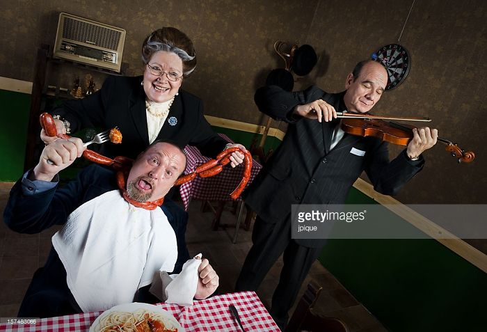 Woman happily strangling a man with sausage while another man is playing the violin