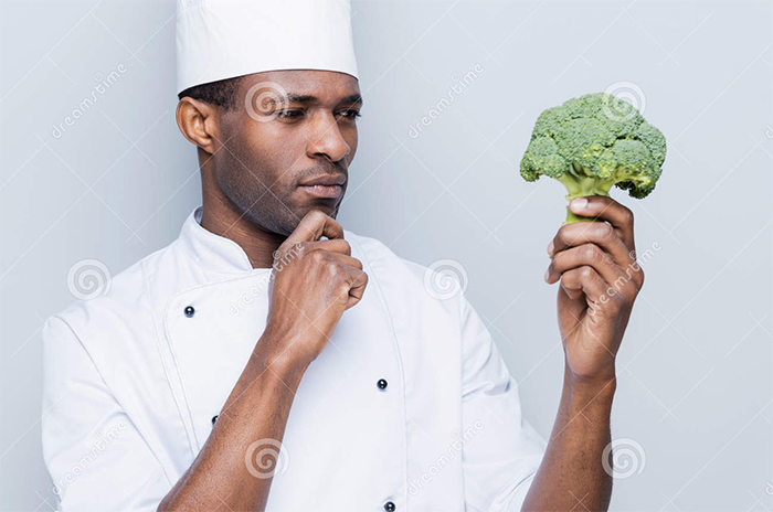 A black man - cooking chef holding the broccoli in his hand and looks very puzzled about it