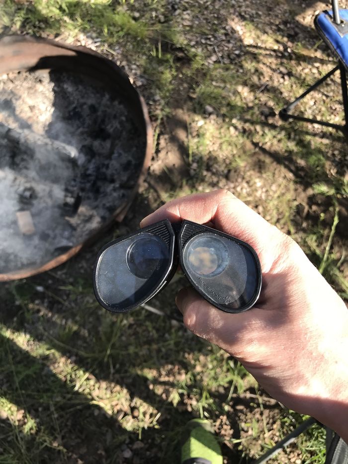 A hand holding sunglasses, unintentionally resembling cosplay goggles, near a campfire in a grassy area.