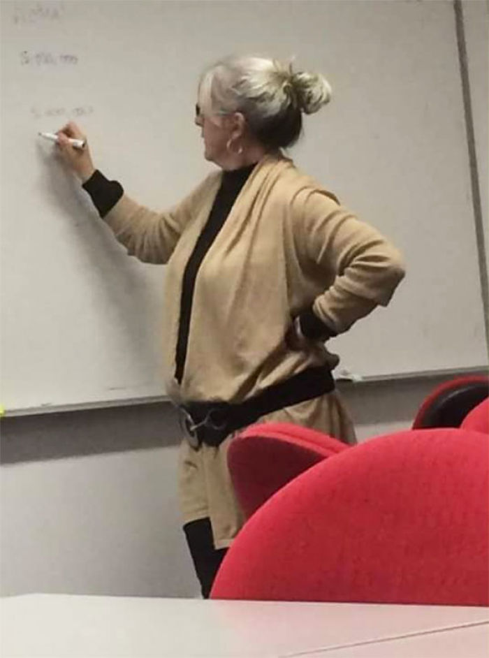 Woman in beige outfit unintentionally resembling a cosplay character, writing on a whiteboard in a classroom setting.