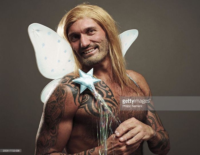 Bare-chested man with tattoos and long hair wears butterfly wings and holds a fairy stick