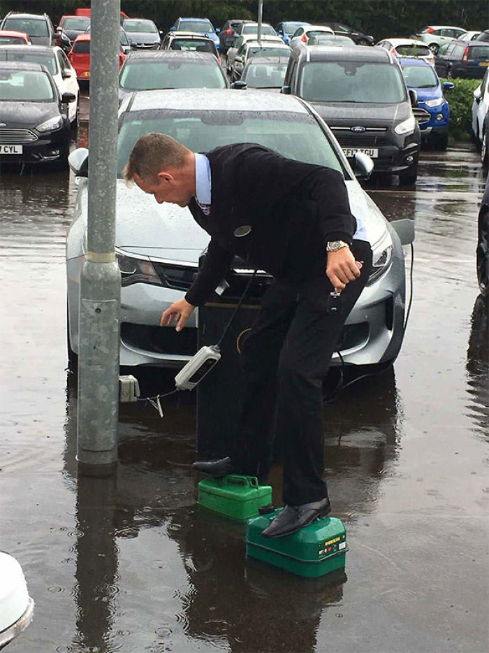 Desenchufando un coche eléctrico mientras usa latas de gasolina para evitar mojarse en la inundación.