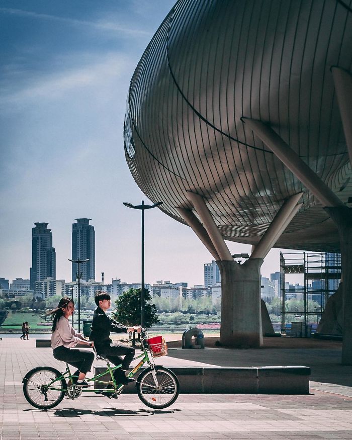 Two people riding a tandem bicycle near modern architecture in Seoul, capturing life as an English teacher in Korea.