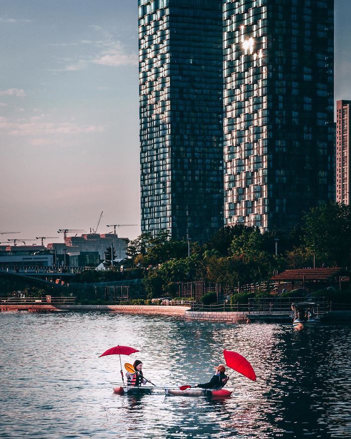 Two people with red umbrellas paddling a kayak near high-rise buildings in Seoul cityscape, reflecting urban life and nature.