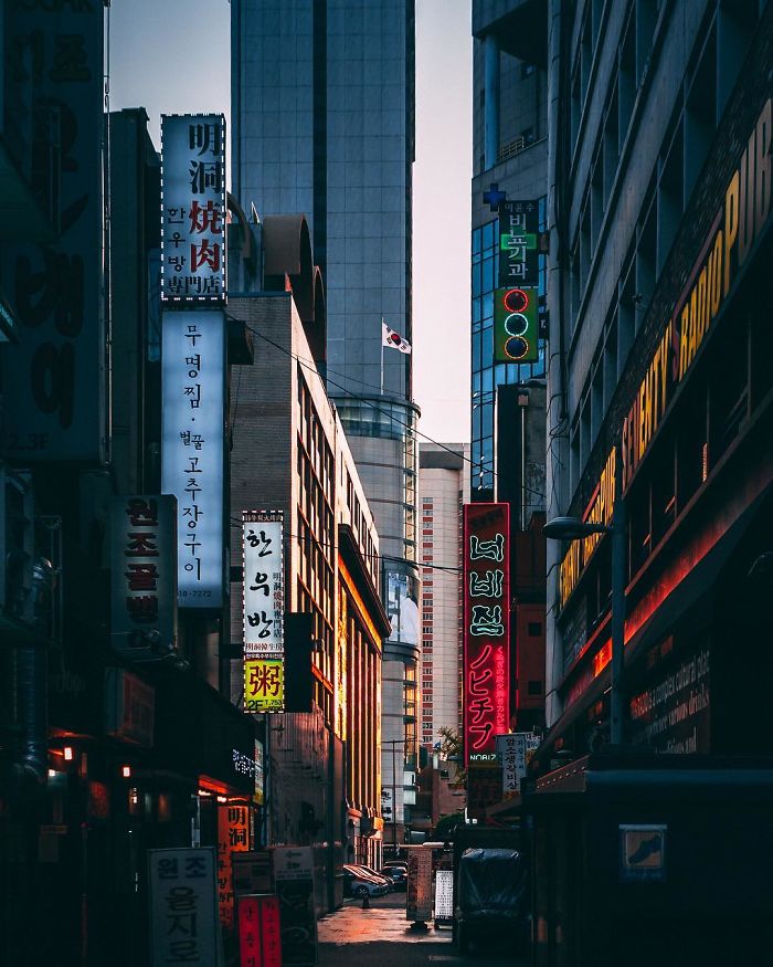 Narrow street in Seoul lined with tall buildings and colorful Korean signs during early evening light.