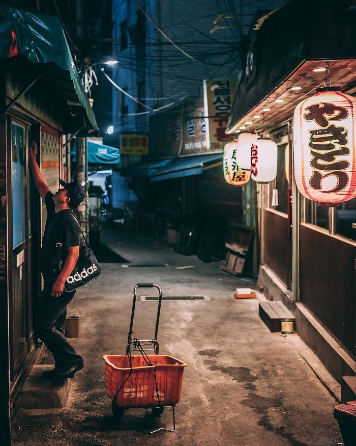 Man with Adidas bag reaching for door in a narrow, dimly lit Seoul alley with hanging lanterns at night.