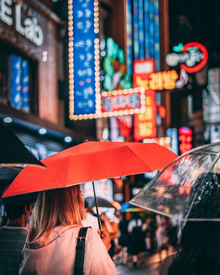 Person holding red umbrella on rainy Seoul street at night with colorful neon signs and busy crowd in the background.