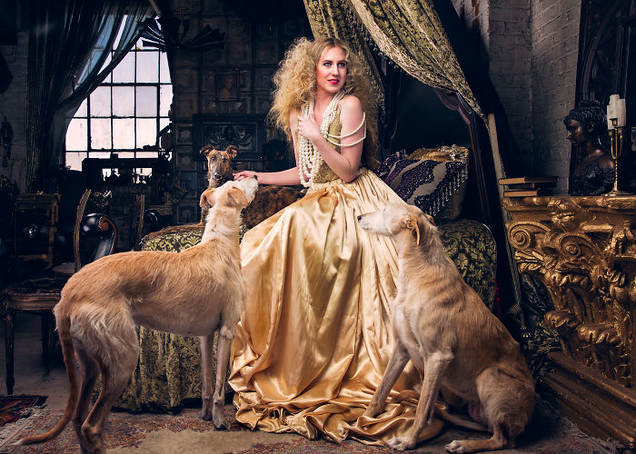 Woman in elegant golden dress sitting with three abandoned hunting dogs in an ornate, vintage room in Spain.
