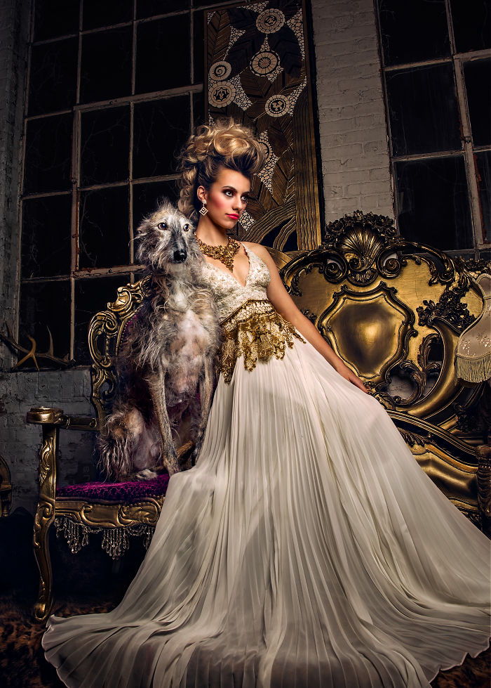 Elegant woman in a detailed gold and white gown posed with an abandoned hunting dog in a vintage ornate setting in Spain.