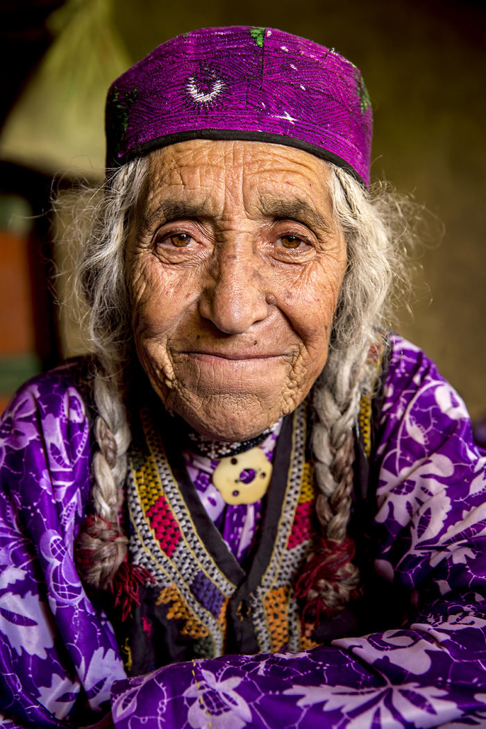 Elderly indigenous woman with braided gray hair wearing colorful traditional clothing and a purple headscarf.