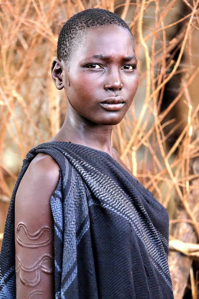 Portrait of an indigenous person with traditional scarification, wearing a dark patterned cloth in a natural setting.