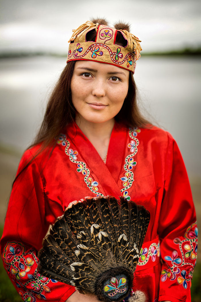 Indigenous woman wearing traditional embroidered red clothing and holding a feathered fan in an outdoor setting.