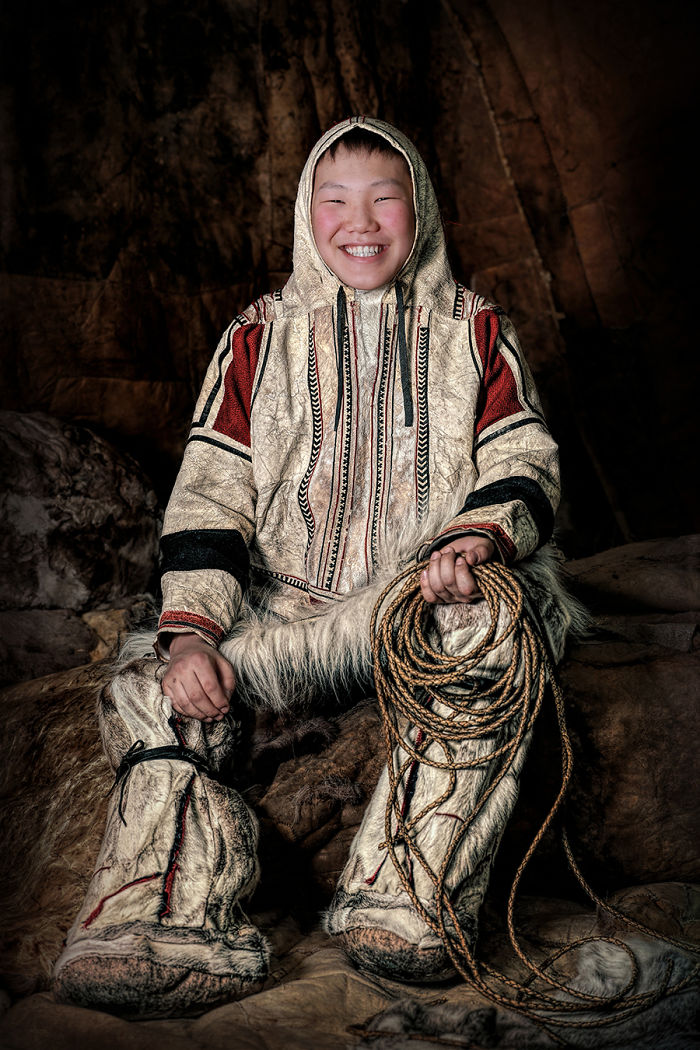 Indigenous person smiling, wearing traditional fur clothing and holding a rope in a dimly lit setting.
