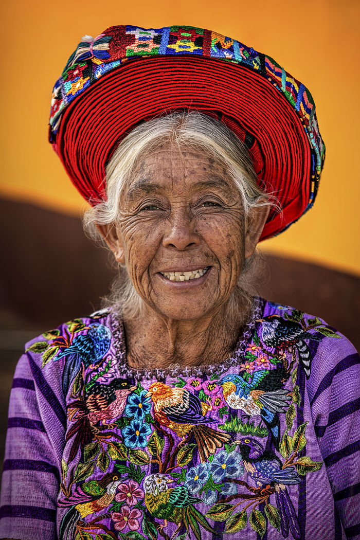 Elderly indigenous woman smiling, wearing a vibrant traditional embroidered blouse and colorful woven hat.