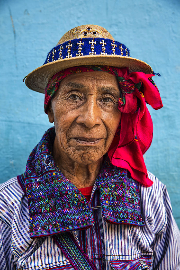 Elderly indigenous man wearing traditional colorful clothing and a straw hat against a blue background.