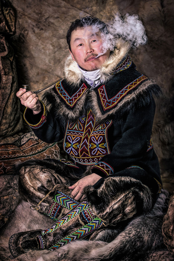 Indigenous man in traditional fur clothing smoking a pipe, captured while traveling to inaccessible corners of the world.