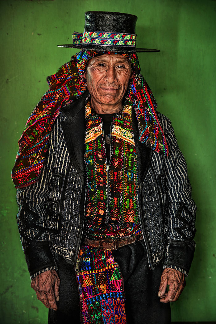 Indigenous man wearing traditional colorful woven clothing and patterned hat standing against a green background.
