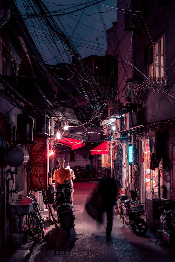 Narrow Shikumen lane house street in Shanghai at night with red awnings, mannequins, bicycles, and tangled overhead wires.