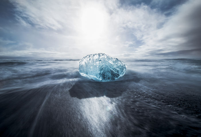 Glowing blue ice chunk on black sand beach with waves and cloudy sky, showcasing Iceland's natural beauty.