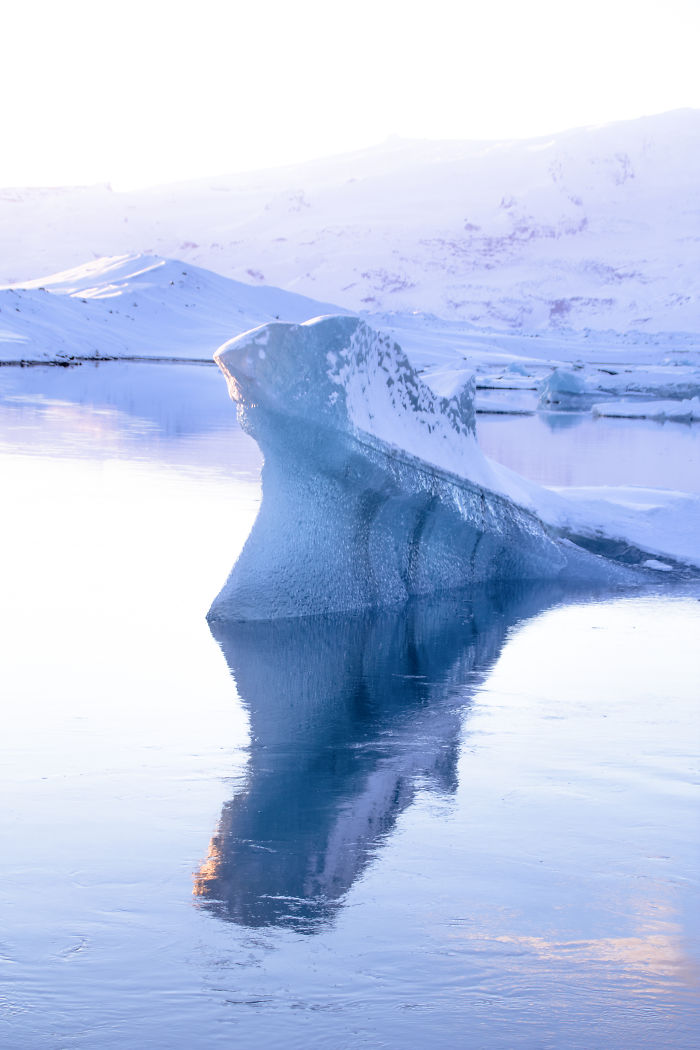 Blue iceberg floating in calm icy waters with snow-covered mountains in the background, showcasing Iceland's natural beauty.