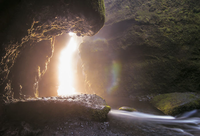 Sunlight shining through a rocky cave opening with moss-covered walls and a flowing stream in Iceland nature.