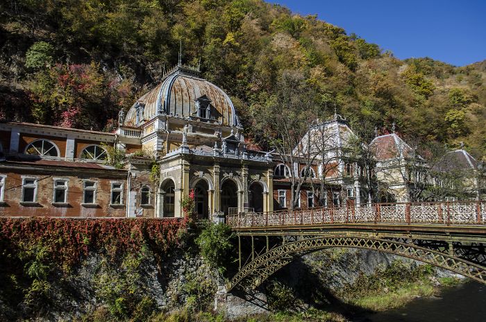 Abandoned thermal baths in Romania surrounded by forested hills and featuring historic architecture and a metal bridge.