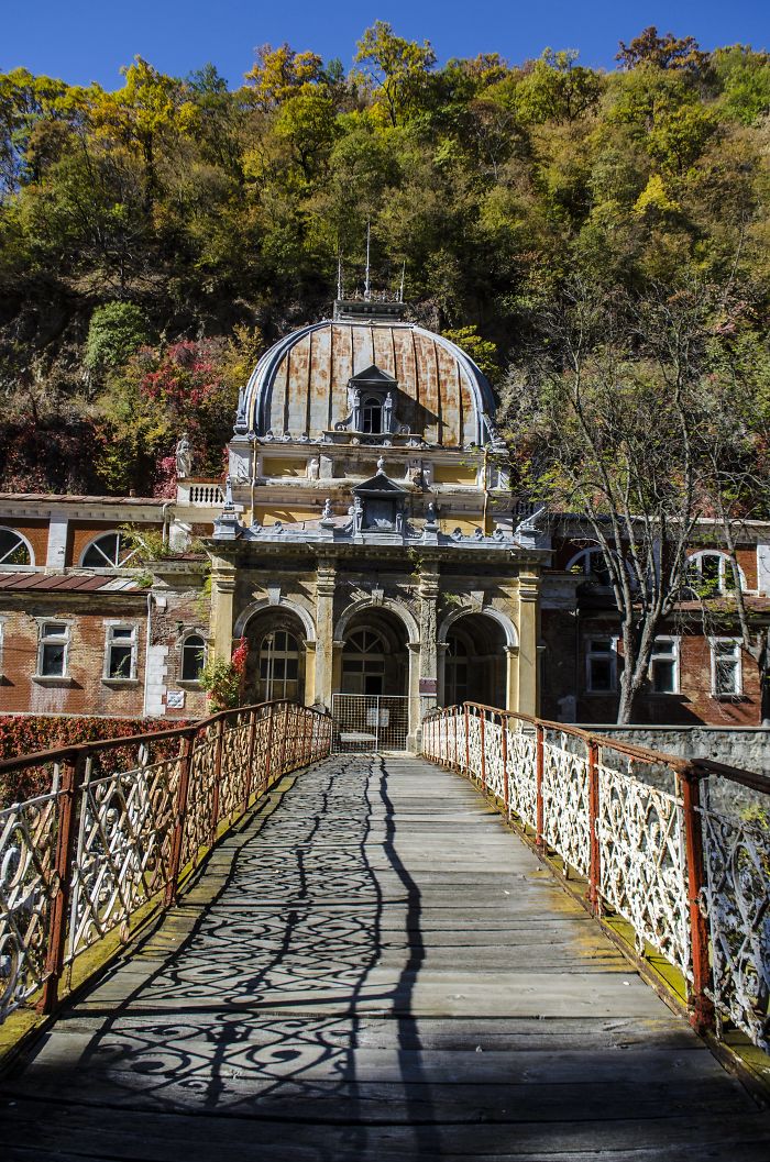 Abandoned thermal baths in Romania with historic entrance and ornate iron bridge surrounded by autumn forest.