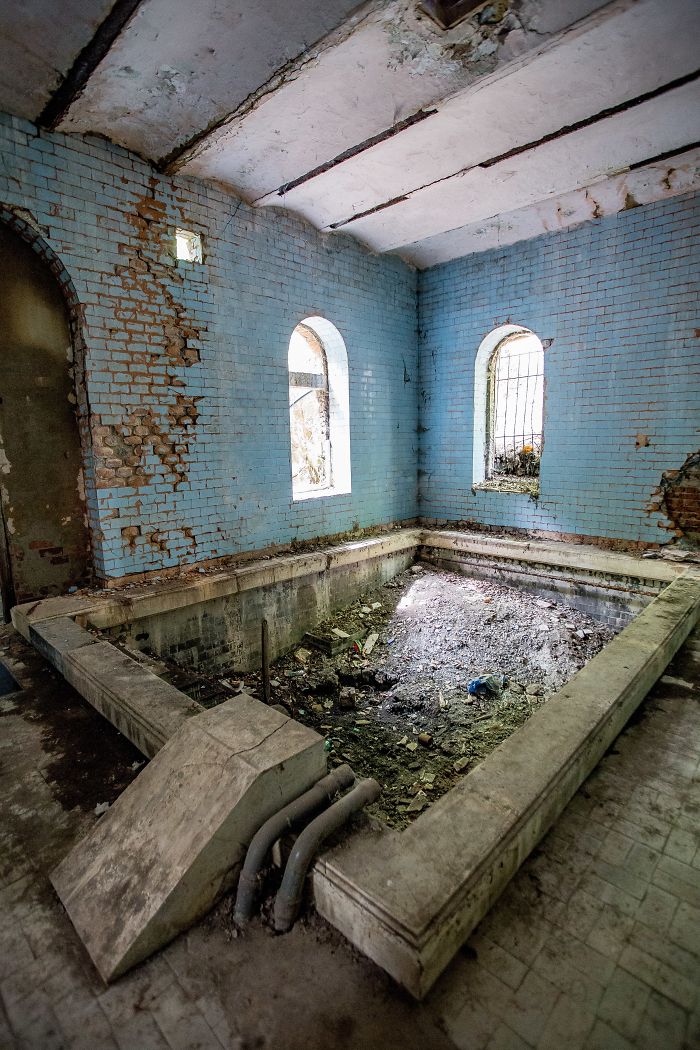 Abandoned thermal baths in Romania showing a damaged empty pool inside a deteriorating blue-tiled room with barred windows.
