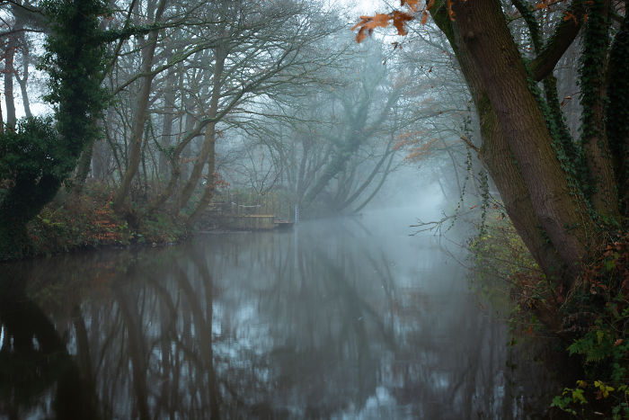 Misty scene of a calm river in the scenic countryside of a rural area in the Netherlands with trees reflected in the water.