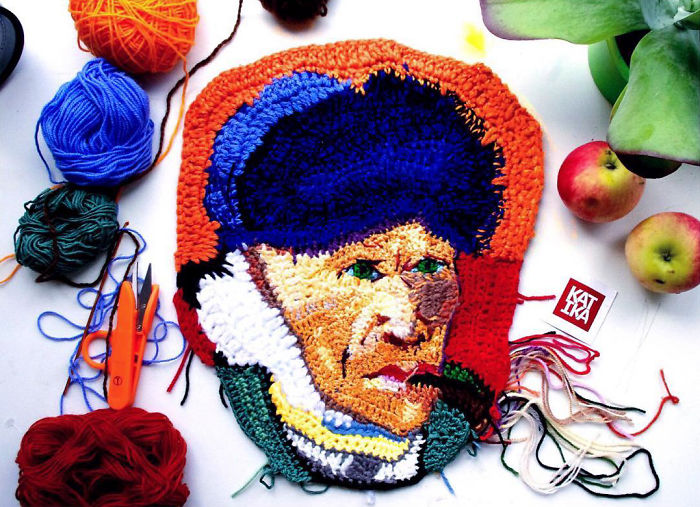 Crochet artist's colorful handmade portrait of a man with a hat, surrounded by yarn and crochet tools on a white surface.