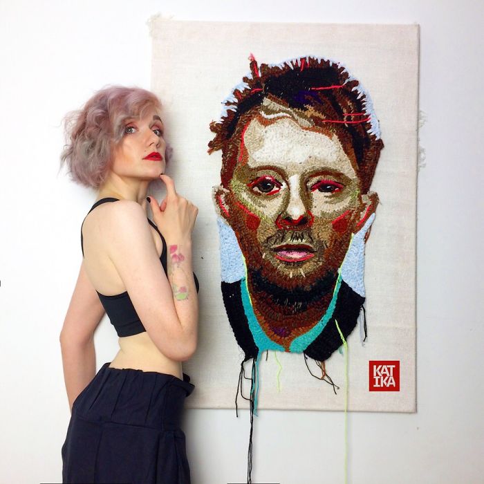 Crochet artist posing beside a detailed crochet portrait with vibrant colors and textured stitching on fabric canvas.