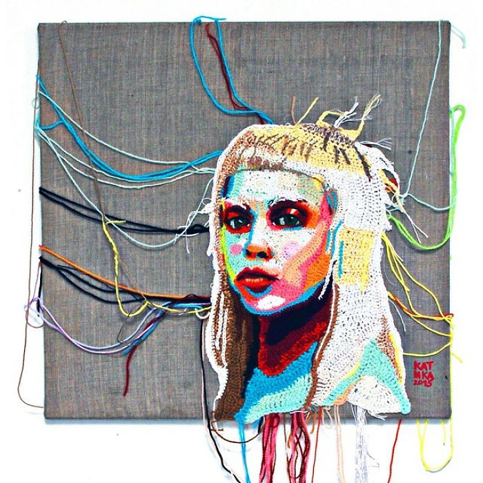 Crochet portrait art of a colorful female face with loose yarn threads on a gray fabric background.