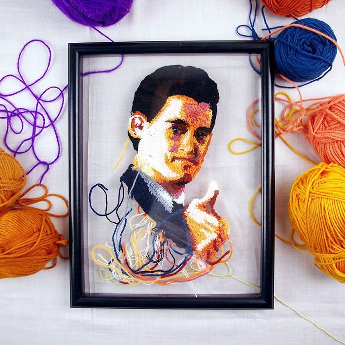 Crochet artist portrait of a man giving a thumbs up, framed and surrounded by colorful yarn balls on a white surface.