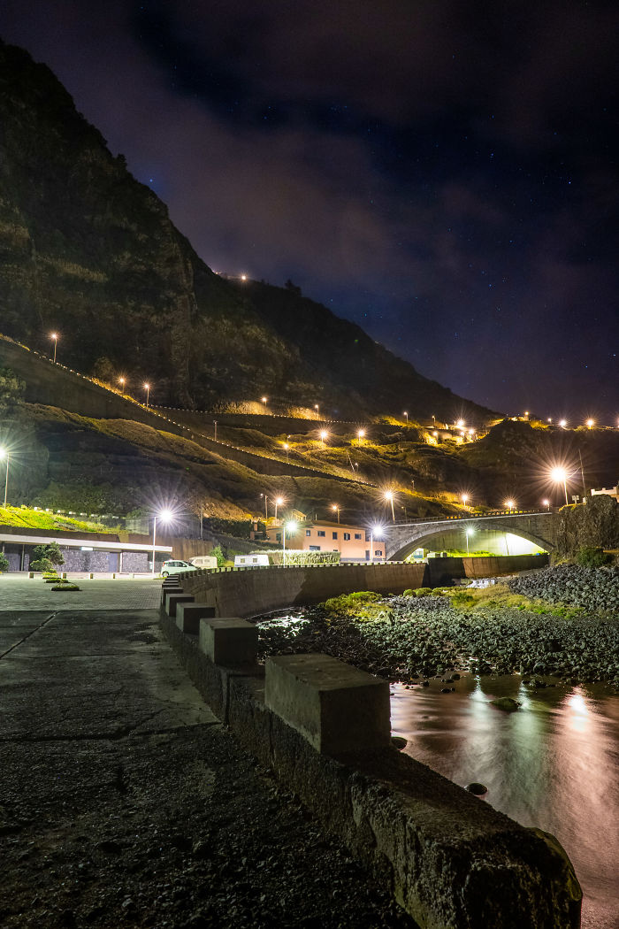 Nighttime view of Madeira coast with illuminated hillside roads, a bridge, and a calm water reflection under a starry sky.