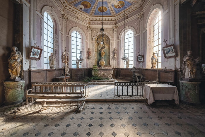 Interior of an abandoned church in Europe featuring statues, tall arched windows, and an ornate altar bathed in natural light.