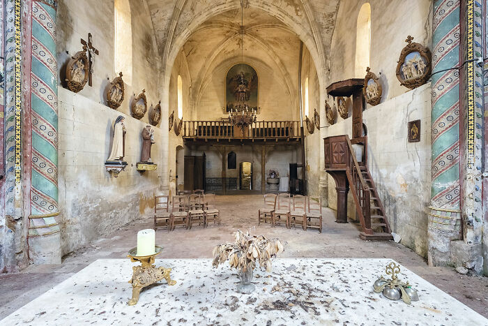 Interior of an abandoned church in Europe showing old chairs, faded religious statues, and a decayed altar with dried flowers.