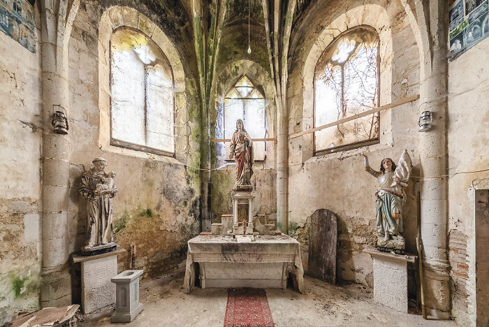 Interior of an abandoned church in Europe with stained glass windows, religious statues, and a weathered altar covered in dust.