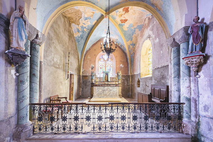 Interior of an abandoned church in Europe with peeling paint, stained glass windows, and religious statues near the altar.