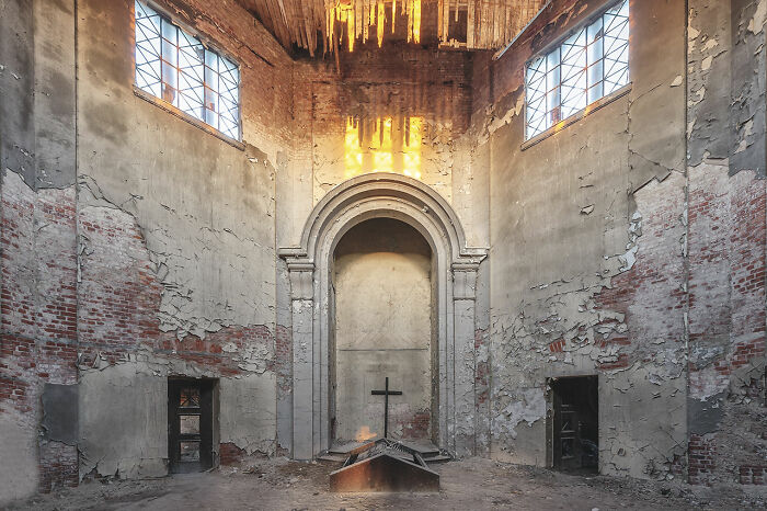 Interior of an abandoned church in Europe with peeling walls, broken windows, and a cross at the altar, showcasing decay.