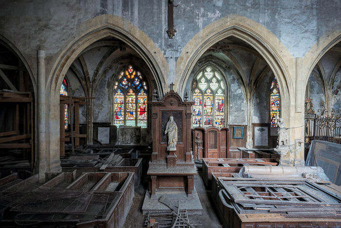 Interior of an abandoned church in Europe featuring stained glass windows, statues, and decaying wooden pews and structures.