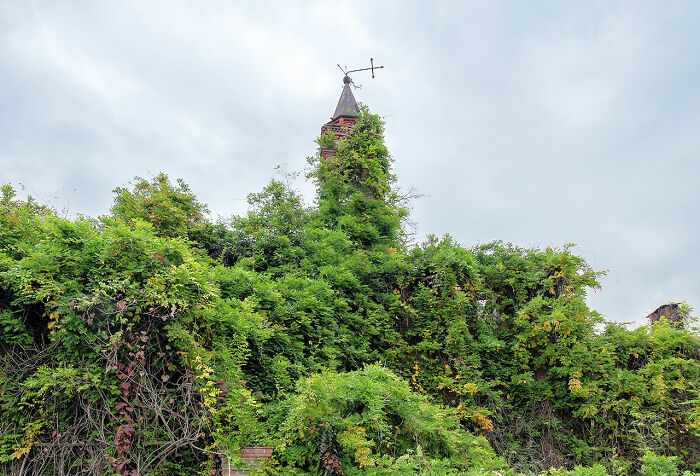Overgrown abandoned church tower covered in green vines and foliage under a cloudy sky across Europe.