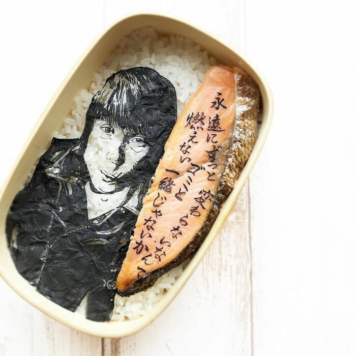 Bento artist creates detailed lunchbox with rice, seaweed portrait, and grilled fish featuring Japanese text.