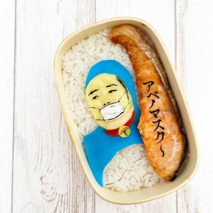Bento artist creates detailed lunchbox art featuring a character wearing a mask alongside a grilled fish piece on rice.